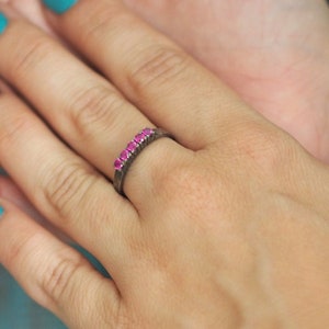 May include: A hand with teal blue nail polish is wearing a silver ring with pink gemstones. The ring is a simple band with a row of small pink stones.