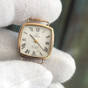 Puede incluir: Un reloj Omega De Ville Quartz dorado con una esfera cuadrada y marcadores de hora en números romanos.
