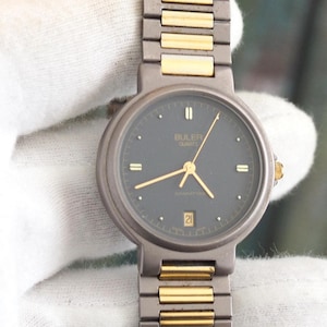 Könnte beinhalten: Eine Armbanduhr mit schwarzem Zifferblatt und goldenen Akzenten an Band und Zifferblatt. Die Uhr hat ein Datumsfenster und die Aufschrift "BULOVA GENEVA" auf dem Zifferblatt.