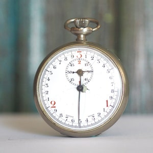 May include: A vintage silver stopwatch with a white face and black hands. Red numerals mark the hours, with a smaller dial at the top. The stopwatch is set against a blurred teal and white background.