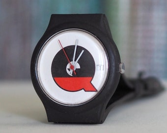 May include: A black wristwatch with a white face and a red and black design. The watch face has a red Q on it.