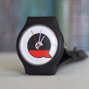 May include: A black wristwatch with a white face and a red and black design. The watch face has a red Q on it.