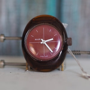 Könnte beinhalten: Eine vintage Armbanduhr aus braunem Kunststoff mit rotem Zifferblatt und weißen Zeigern. Auf dem Zifferblatt der Uhr steht "BULER".