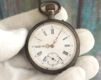 Puede incluir: Un reloj de bolsillo de plata con números romanos en la esfera. El reloj tiene una esfera blanca con agujas negras y una pequeña aguja de segundos.