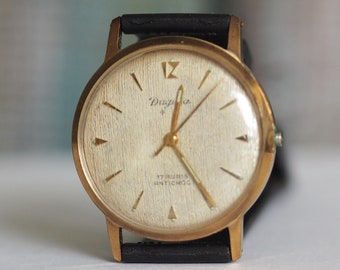 Puede incluir: Un reloj de pulsera dorado con una esfera blanca y una correa de cuero negra. La esfera del reloj tiene marcadores de hora en números romanos negros y una pequeña estrella sobre la palabra "Dugena" en la parte superior.