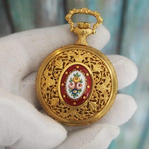 May include: Ornate gold-tone pocket watch with a floral design. The watch features an oval, red enamel center with a painted floral bouquet. The watch has a decorative filigree pattern and a loop at the top.