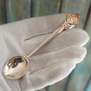 May include: A silver teaspoon with a decorative handle featuring a raised design of two people's profiles. The handle is shaped like a diamond and has a small crown on top.