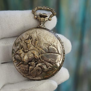 May include: Antique silver pocket watch with a detailed relief of deer in a forest scene. The watch has a decorative handle and a textured, aged appearance, suggesting it is a vintage item. The watch is round and held by a gloved hand.