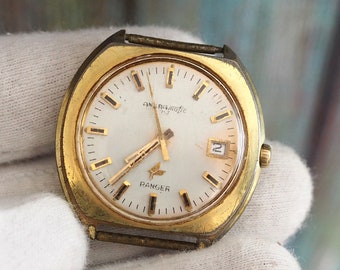 Puede incluir: Un reloj vintage dorado con esfera blanca. La esfera muestra el texto "ANBAZOTE" y "RANGER", junto con la fecha. El reloj tiene manecillas e indicadores de hora dorados. El reloj está sujeto por un guante blanco.