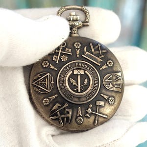 May include: Antique silver-toned pocket watch with intricate embossed detailing. The watch face features various symbols and the text "SEGNE DAS EHRBARE HANDWERK". A decorative loop and crown are at the top. The watch is held in a white glove.