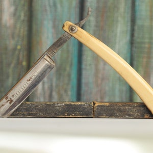 May include: A vintage straight-edge razor with a bone handle and a metal blade. The blade is inscribed with the text "GOODPLEX" and "MADE IN GERMANY". The razor is resting on a wooden box.
