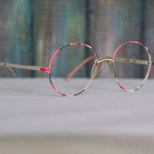 May include: Round eyeglasses with a silver frame and a pink, blue and white patterned rim.