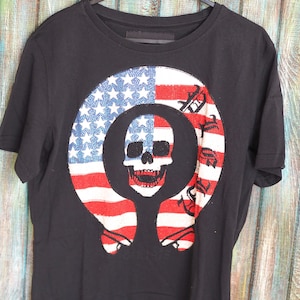 Puede incluir: Camiseta negra con un gráfico de una calavera dentro de un círculo con un diseño de bandera estadounidense. El texto "Ride Free" está impreso alrededor del círculo.
