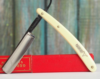 May include: A vintage straight razor with a white plastic handle and a silver blade. The razor is resting on a red box with gold lettering that reads "GEB. SOL. N."