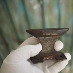 Puede incluir: Un pequeño yunque de metal oxidado con una parte superior plana y dos agujeros en la parte inferior. El yunque se sostiene en una mano enguantada.