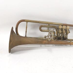 May include: An antique brass flugelhorn with a curved design. The instrument features a flared bell, valves, and a mouthpiece. The flugelhorn has a tarnished, aged appearance, suggesting it is vintage. The instrument is a wind instrument.