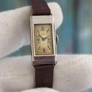 May include: A vintage rectangular wristwatch with a silver-tone case and a brown leather band. The watch face is cream-colored with black numerals and hands. The watch is approximately 0.75 inches wide and 1.5 inches long.