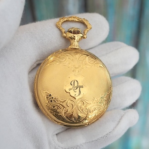 May include: A gold pocket watch with ornate floral engravings and a shield-shaped monogram. The watch has a decorative loop at the top and a textured surface. The watch is a classic timepiece.