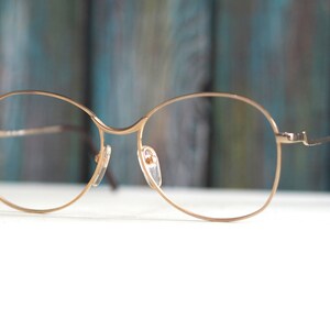 May include: Gold-tone metal spectacles with round lenses. The glasses are on a white surface.