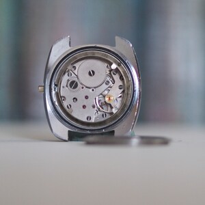 May include: Close-up of a vintage watch movement, showcasing intricate gears, jewels, and mechanical components within a silver-colored case. The watch's internal mechanisms are visible, highlighting the precision of the craftsmanship.