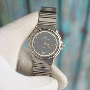 Puede incluir: Un reloj de cuarzo Maurice Lacroix plateado con esfera gris oscuro. El reloj tiene una pantalla de fecha a las 6 en punto y una correa plateada. El reloj está sujeto por una mano enguantada.