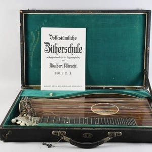 Può includere: Una cetra vintage in una custodia nera con interno in velluto verde. Lo strumento ha numerose corde e un corpo in legno. Un foglio di carta con testo tedesco è all'interno della custodia. La custodia è aperta, rivelando lo strumento.