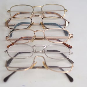 May include: Five pairs of spectacles stacked on top of each other. The spectacles are all different styles and colours, with some having gold frames and others having silver frames.