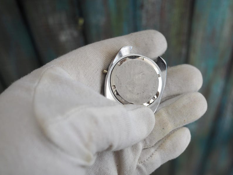 May include: A silver-toned watch case with a round, textured face. The watch is held in a white gloved hand. The watch face has small text and markings.