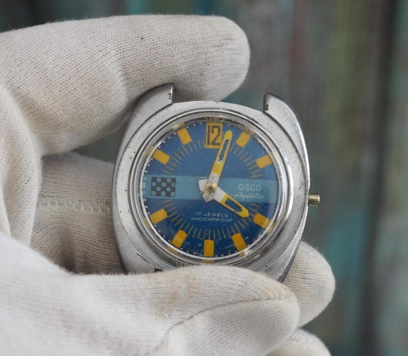 May include: Vintage OSCO Aquatica wristwatch with a blue and yellow face. The watch has a silver-tone case and features the text "17 Jewels Shockproof". The watch is held by a gloved hand.