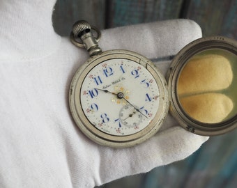 Puede incluir: Un reloj de bolsillo de plata con una esfera blanca y marcadores de hora romanos azules. El reloj tiene un diseño floral dorado en el centro y las palabras "Solar Watch Co." impresas en la esfera.