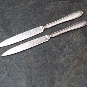 May include: Two silver butter knives laid out on a dark gray textured surface.