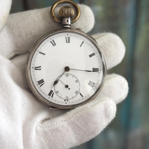 May include: A silver pocket watch with a white face and black Roman numerals. The watch has a smaller subdial and a gold-colored crown. The watch is held by a gloved hand.