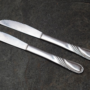 Op de afbeelding: Twee zilveren botermesjes met sierlijke handvatten. De messen hebben een gekarteld lemmet en liggen op een donkergrijs, getextureerd oppervlak.