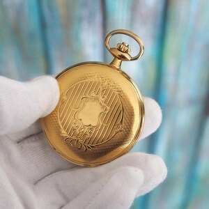 May include: A gold pocket watch with an ornate, engraved design. The watch features a detailed shield-like emblem surrounded by floral patterns and vertical lines. The watch is held in a white gloved hand against a blurred teal and blue background.