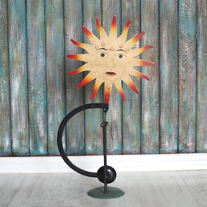 Può includere: Una scultura solare in metallo stravagante con finitura anticata. Il sole ha un volto con occhi e bocca, con raggi rossi e gialli. La scultura è montata su un supporto in metallo nero con un design circolare.