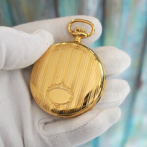 May include: A gold pocket watch with a textured, striped design on the cover. The watch has a small, oval-shaped decorative element and a loop at the top. The watch is held in a white glove.