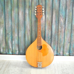 Puede incluir: Un mandolín de madera vintage con un acabado marrón claro y un agujero de sonido con una roseta decorativa. El mandolín tiene un diapasón de madera y un juego de clavijas de afinación en la pala.