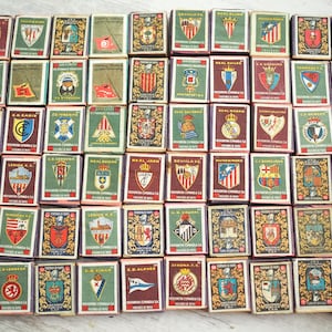 May include: A collection of vintage matchboxes, each featuring a different team emblem. The matchboxes are rectangular, with a variety of colors and designs. The emblems include crests, flags, and team names, suggesting a theme of sports or collectibles.