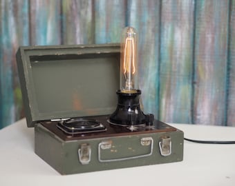 Könnte beinhalten: Eine Lampe im Vintage-Stil mit einer klaren Glühbirne und einem schwarzen Sockel, montiert auf einer grünen Metallbox mit zwei Riegeln und einem Griff.