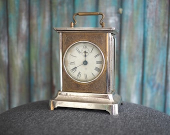 Könnte beinhalten: Antike silber- und goldfarbene Reiseuhr mit weißem Zifferblatt und schwarzen römischen Ziffern. Die Uhr hat einen Griff und steht auf einem silbernen Sockel. Das Zifferblatt hat schwarze Zeiger.