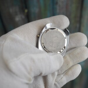 May include: A silver-toned watch case with a round, textured back. The watch is held in a white glove. The watch's back has etched details. The watch is a vintage style.