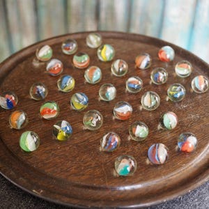 May include: A wooden game board with a circular depression in the center, filled with colorful marbles. The marbles are arranged in a circular pattern, with some marbles overlapping others.