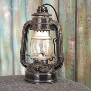 Op de afbeelding: Een zwarte vintage olielamp met een glazen globe en een metalen handvat. De lamp staat op een grijs oppervlak voor een houten achtergrond.