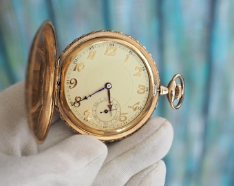 May include: A gold pocket watch with an open lid, showing a cream face with gold numerals and hands. The watch has a small seconds dial. The watch is held in a white gloved hand. The watch case has an ornate design.