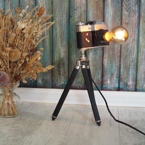 Könnte beinhalten: Eine Vintage-Kamera-Lampe mit einem schwarzen und silbernen Gehäuse, montiert auf einem schwarzen Stativ. Die Kamera hat eine Glühbirne im Objektiv und ein schwarzes Kabel.