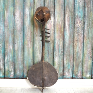 May include: A wooden musical instrument with a round, brown, leather-covered body and a carved wooden head with a face. The instrument is standing upright against a blue and white wood panel background.