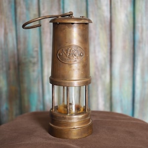 Op de afbeelding: Antieke messing mijnwerkerslamp met een heldere glazen cilinder en een handvat. De lamp heeft een ovaal embleem met een sierlijk ontwerp. De lamp staat op een bruin oppervlak.