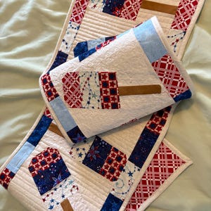 May include: Handmade patriotic quilt with red, white, and blue popsicle designs. The quilt has a white background with star and stripe patterns, and brown wooden popsicle stick accents. The quilt is likely made of cotton fabric.