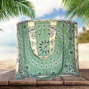 Puede incluir: Un bolso tote grande con un estampado de cachemira verde y blanco. El bolso tiene una correa ancha de color crema y rayas verticales crema. El bolso está hecho de una tela suave y está sobre una superficie de madera.