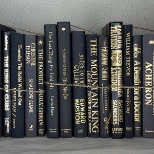 May include: A collection of black hardcover books with gold lettering, arranged on a shelf and bound with twine. Titles include "Nobody Nowhere" and "The Mountain King". The books create a sophisticated, minimalist aesthetic.